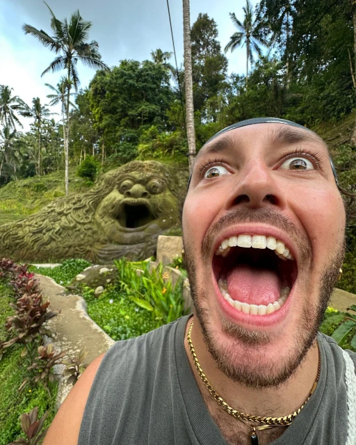 Man with a wide smile and open mouth takes a selfie with a stone sculpture in Bali surrounded by tropical greenery and palm trees.