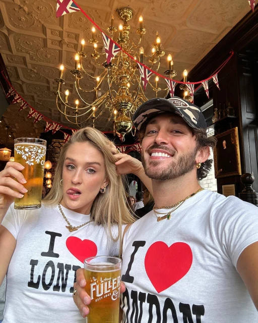 Two people in 'I love London' shirts holding beer glasses in a pub with British flags, beneath a vintage chandelier, smiling.