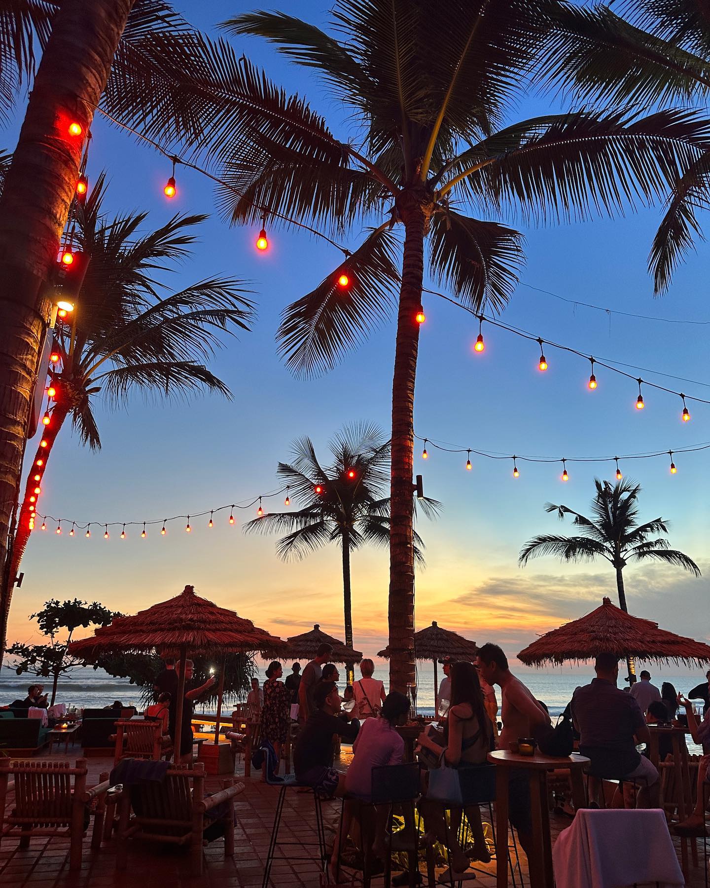 Sunset view of a beachfront resort with palm trees, string lights, thatch umbrellas, tables, and people gathered at dusk.