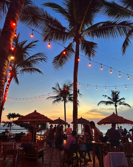 Sunset view of a beachfront resort with palm trees, string lights, thatch umbrellas, tables, and people gathered at dusk.