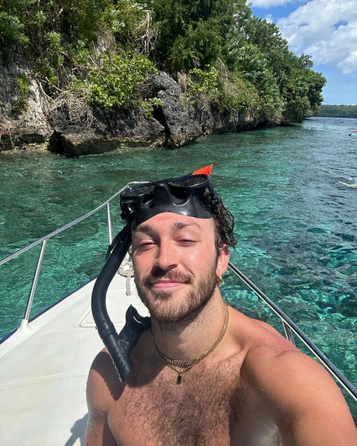 Shirtless man on a boat with snorkel gear smiling at the camera with a rocky tree covered island in the background on a sunny day.