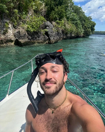 Shirtless man on a boat with snorkel gear smiling at the camera with a rocky tree covered island in the background on a sunny day.