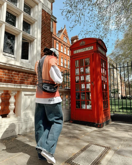 A person stands on a sidewalk next to a red telephone booth in an urban area, with brick buildings and leafy trees visible in the background.