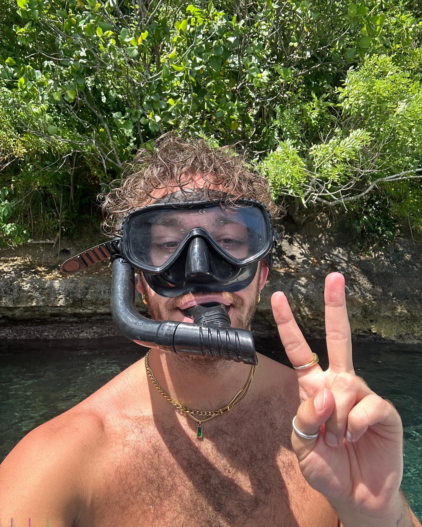 Adult male with curly hair in dive mask makes peace sign outdoors near water and lush green foliage, wearing gold necklace and rings.