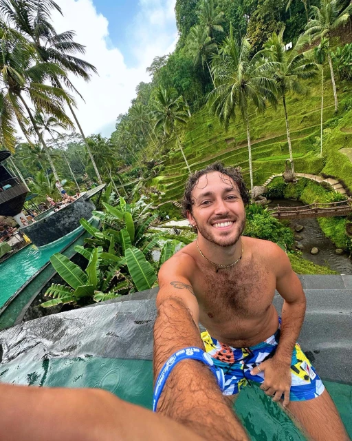 Smiling man in swimwear takes a selfie with tropical rice terraces and palm trees in the background on a sunny day in Bali.