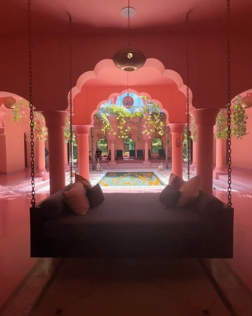 Gray daybed with pillows hangs in the pink interior of a building, overlooking a pool surrounded by plants and chairs.