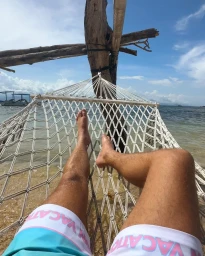 POV shot of a person relaxing in a rope hammock strung between wooden posts above a tropical beach with turquoise water.