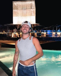 Smiling man in a grey tank top and white cap standing outdoors at night near an illuminated pool and bridge.