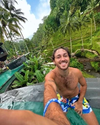A shirtless man smiling while taking a selfie in a pool surrounded by lush tropical greenery and palm trees.