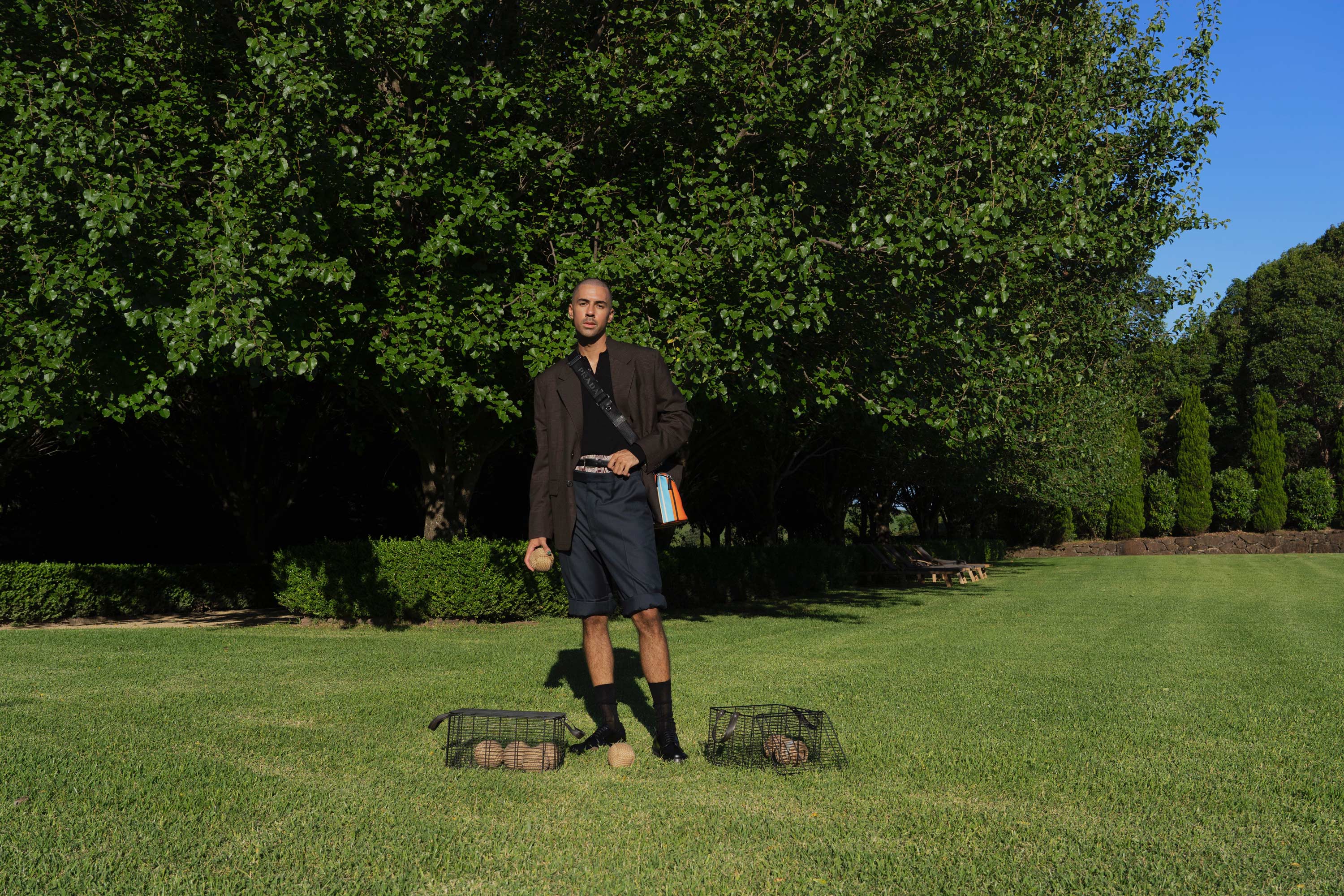 A man standing on a green lawn between two animal traps holding balls, with trees in the background, under a blue sky.