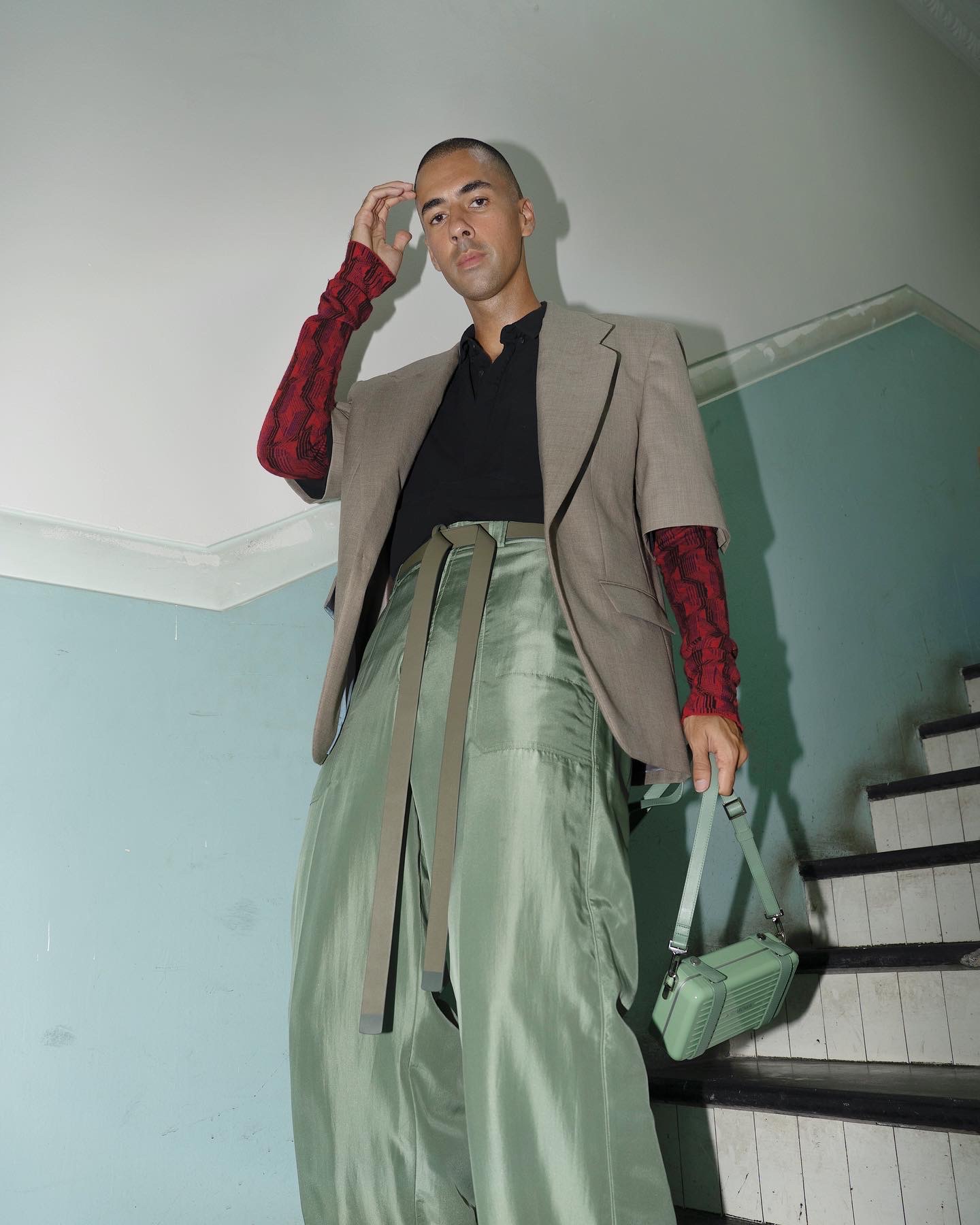 Man in beige blazer and shiny green pants stands on stairs, holding a small boxy handbag. Fashion-forward pose.