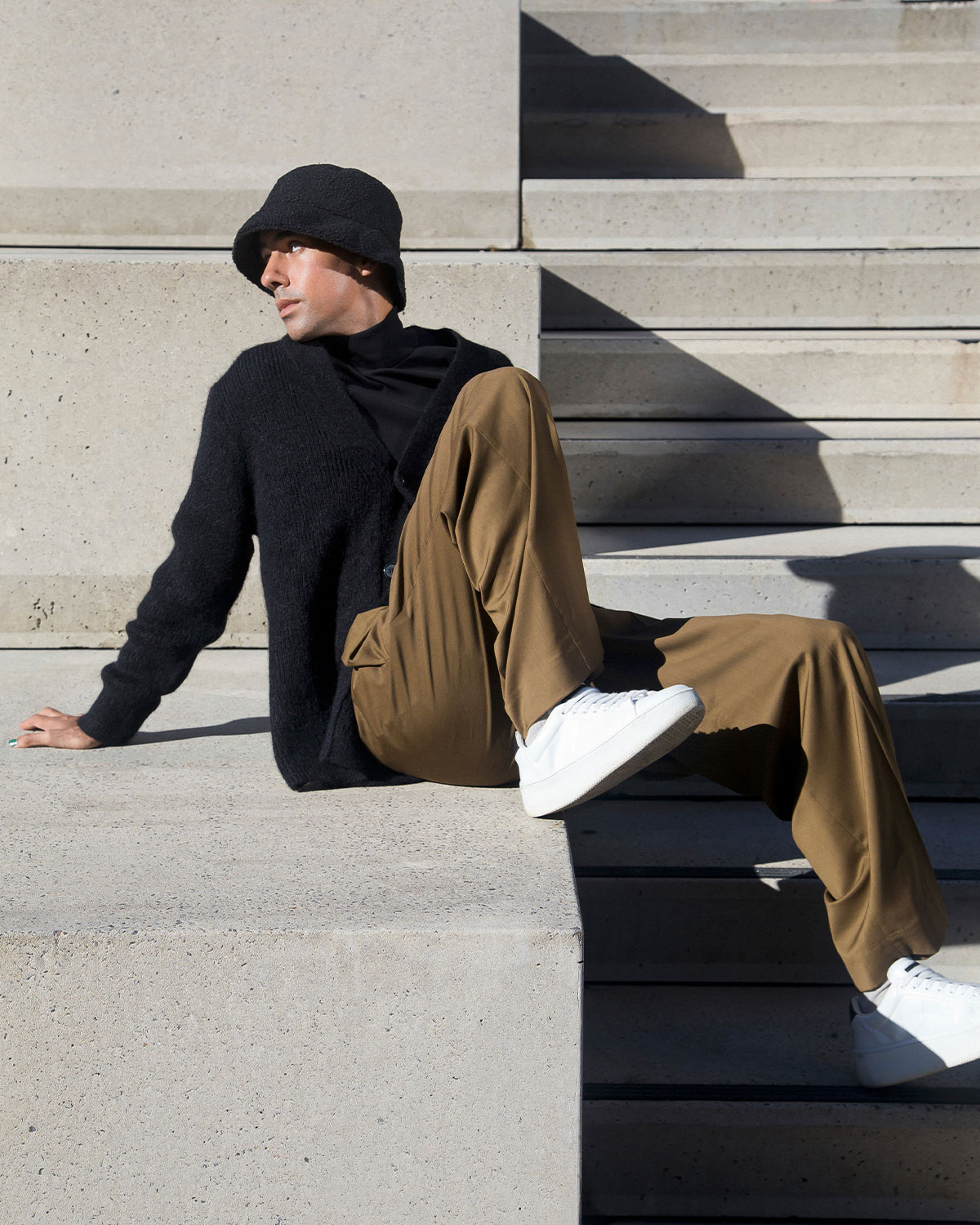 A stylish adult male sits on concrete steps outdoors, dressed in a dark sweater, olive-green pants, white sneakers, and a black bucket hat.