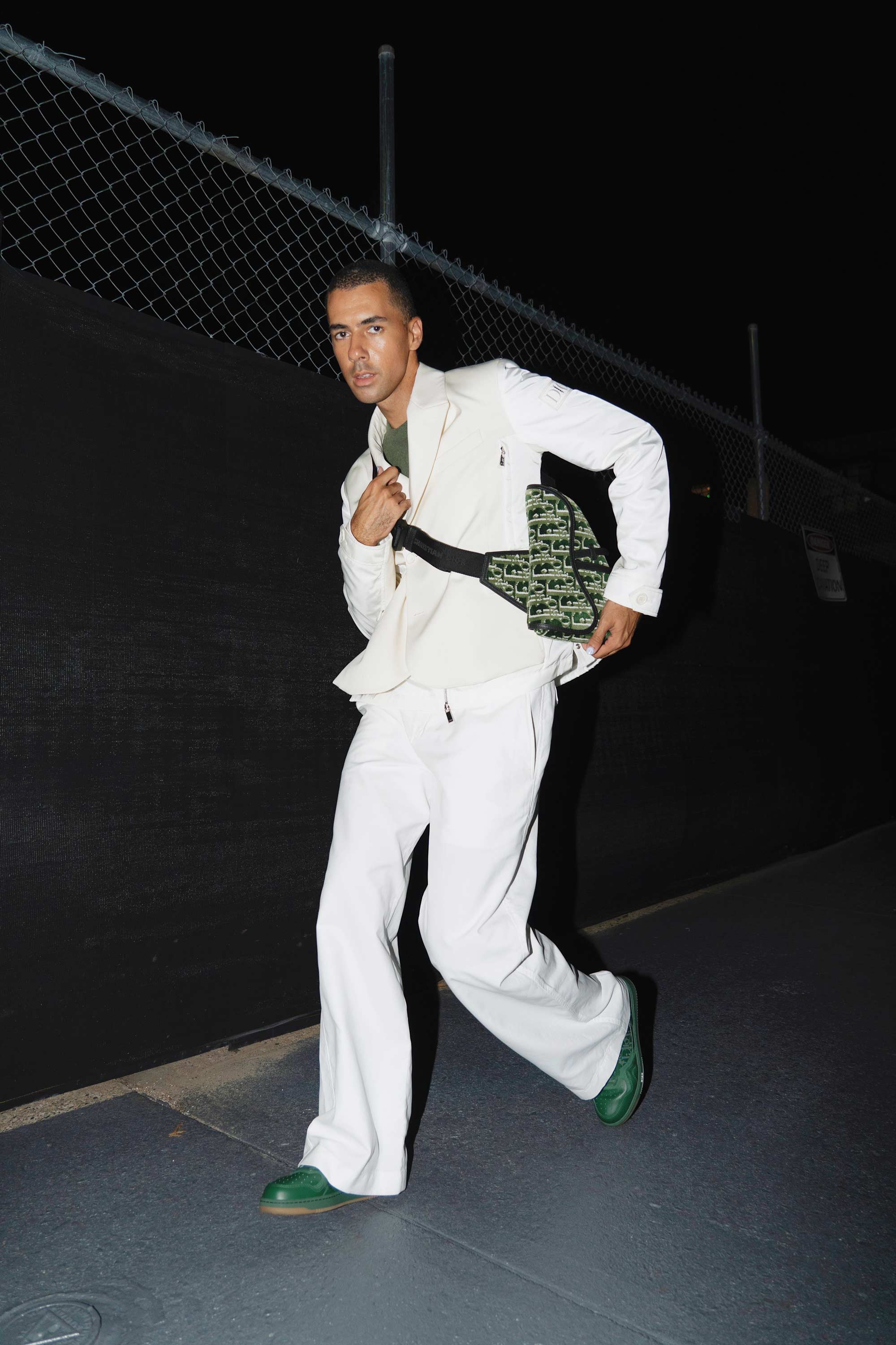A stylish adult male is captured in motion outdoors at night wearing a white outfit with a black waist bag and green shoes.