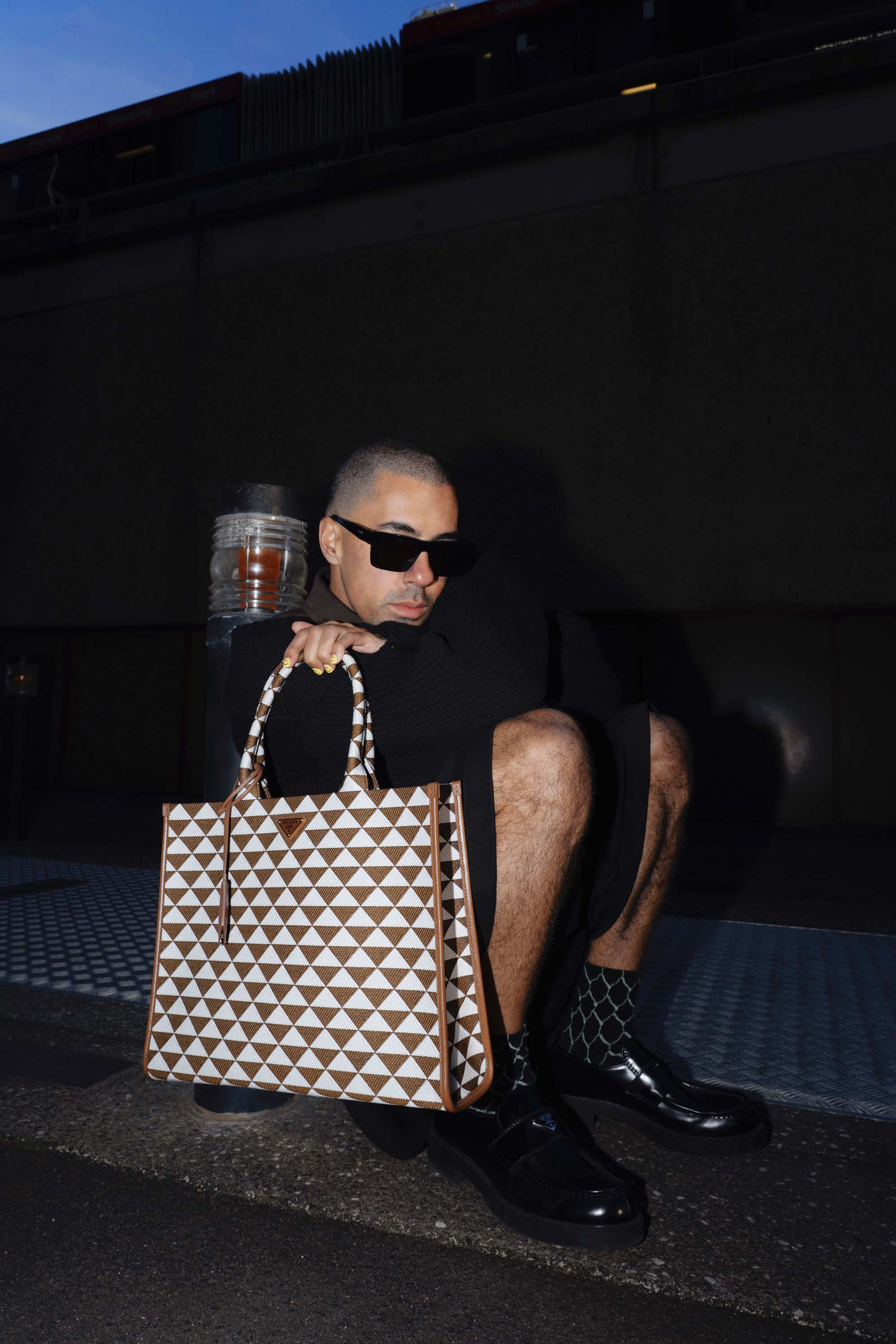 Adult male crouches outdoors holding a designer tote bag, wearing sunglasses and black clothing, with glossy black shoes and patterned socks.