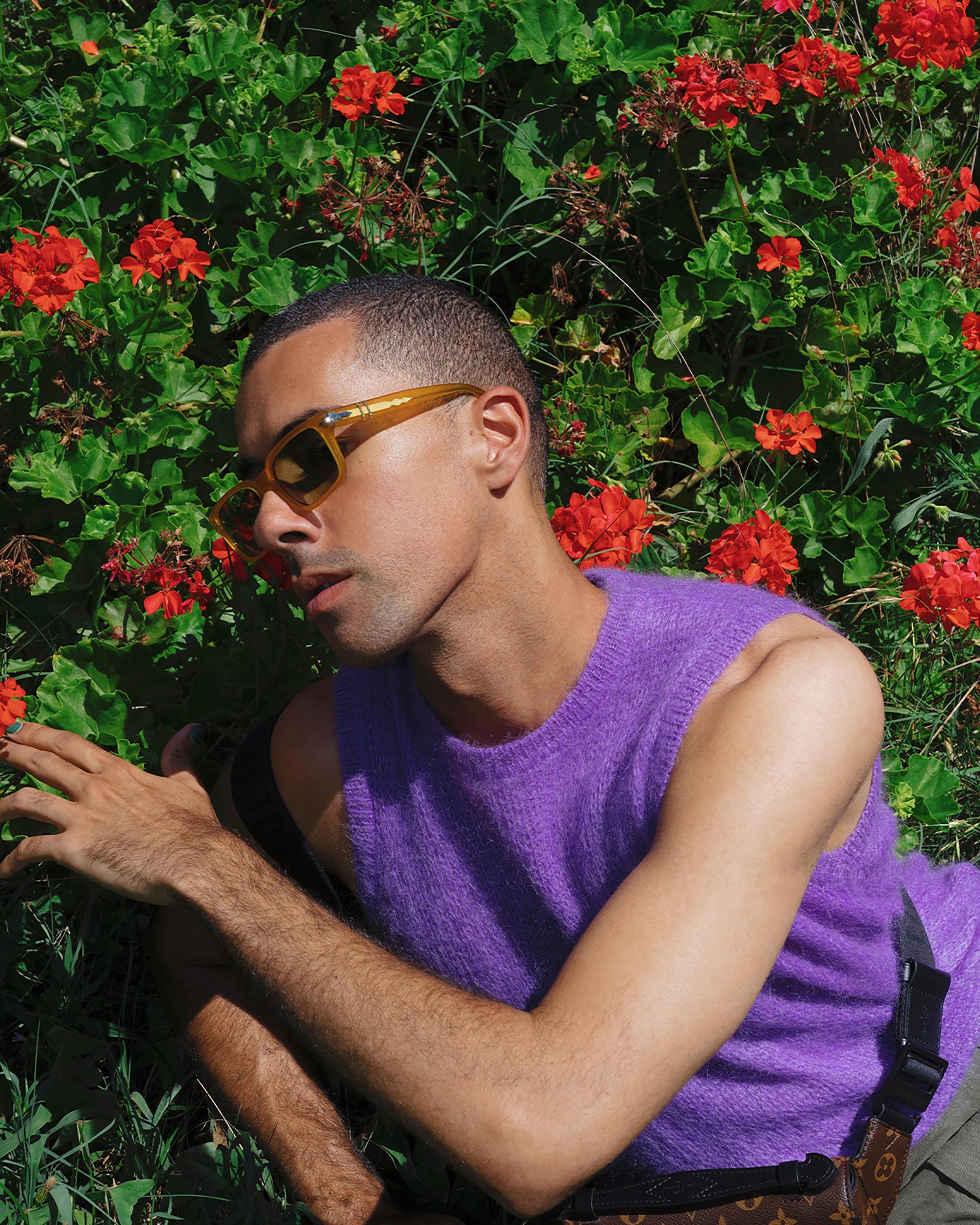A young adult male poses outdoors in a purple sleeveless top and amber sunglasses, surrounded by green foliage and red geraniums.