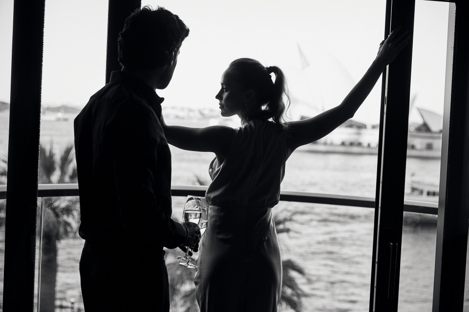 Silhouetted couple enjoys champagne at a window overlooking the Sydney Opera House and harbour, black and white, indoor setting.