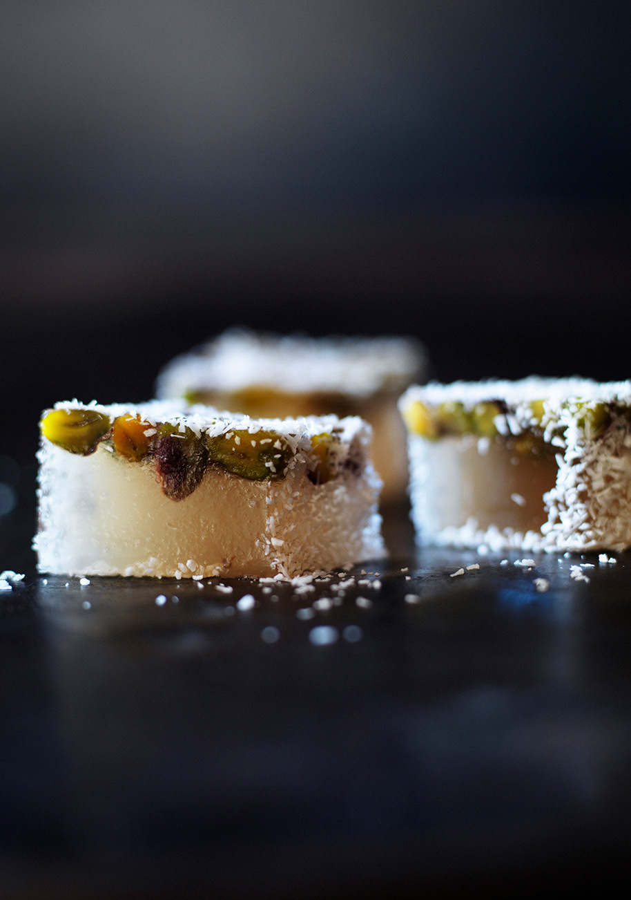 Three pieces of Turkish delight confection topped with nuts and coconut flakes on a dark surface, studio lighting, close-up shot.