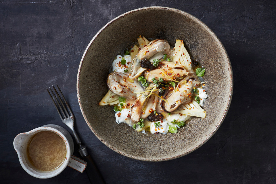 Overhead shot of oyster appetizer with lemon and herbs in stoneware bowl, fork and gravy boat on textured dark gray surface.