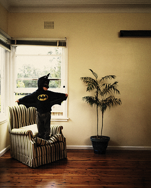 Young child dressed in a Batman costume standing on a striped armchair in an indoor living room with hardwood floors and a window.
