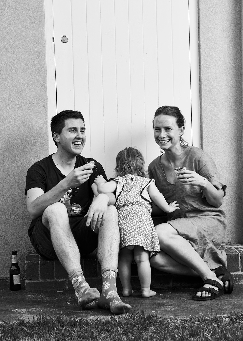 Monochrome photo of a family of three—parents and toddler—sitting outside on a stoop, enjoying snacks and a relaxed atmosphere.