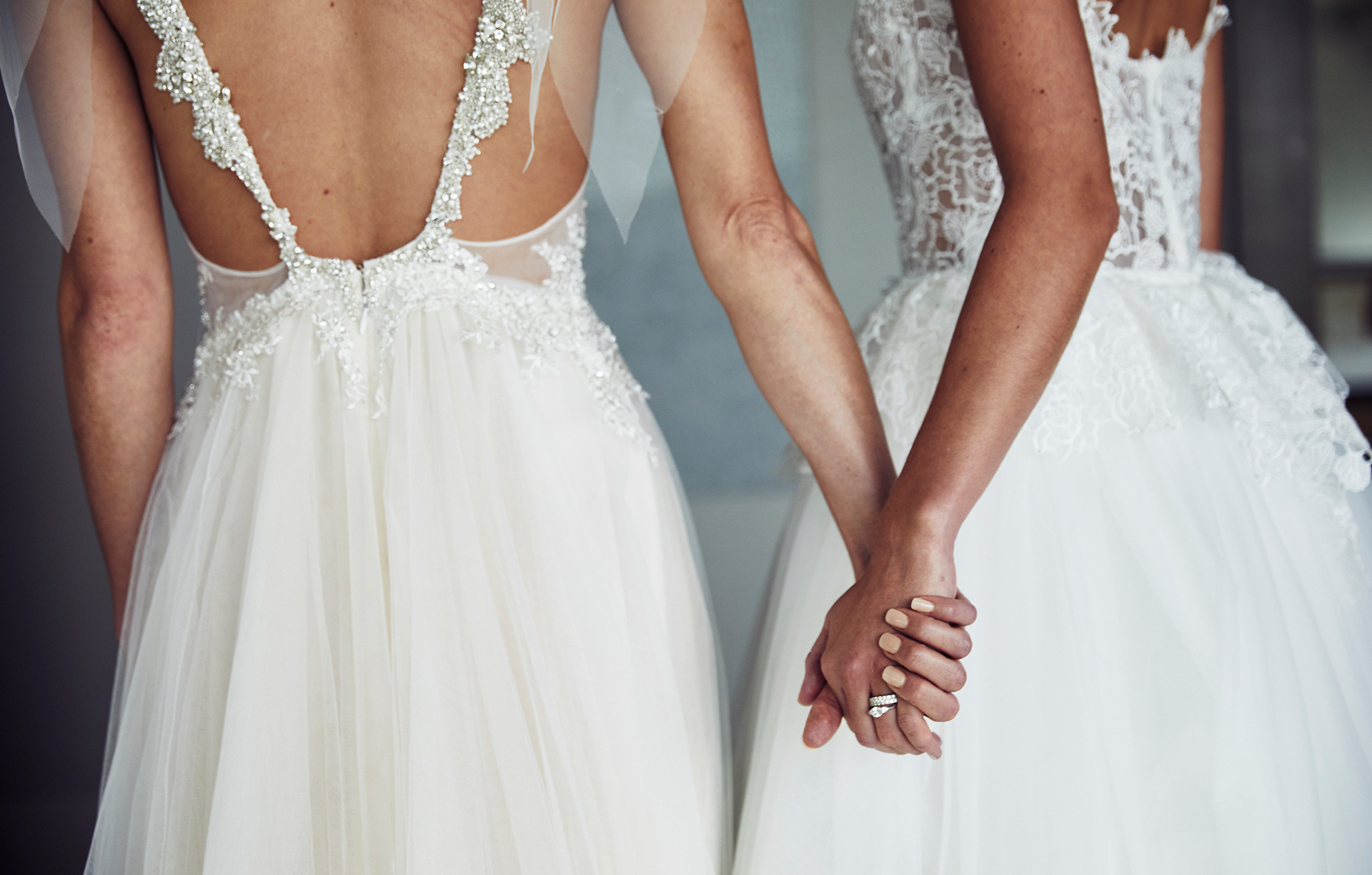 Two brides in white dresses holding hands, showcasing intricate lace details and soft lighting, captured in a close-up shot. Wedding theme.