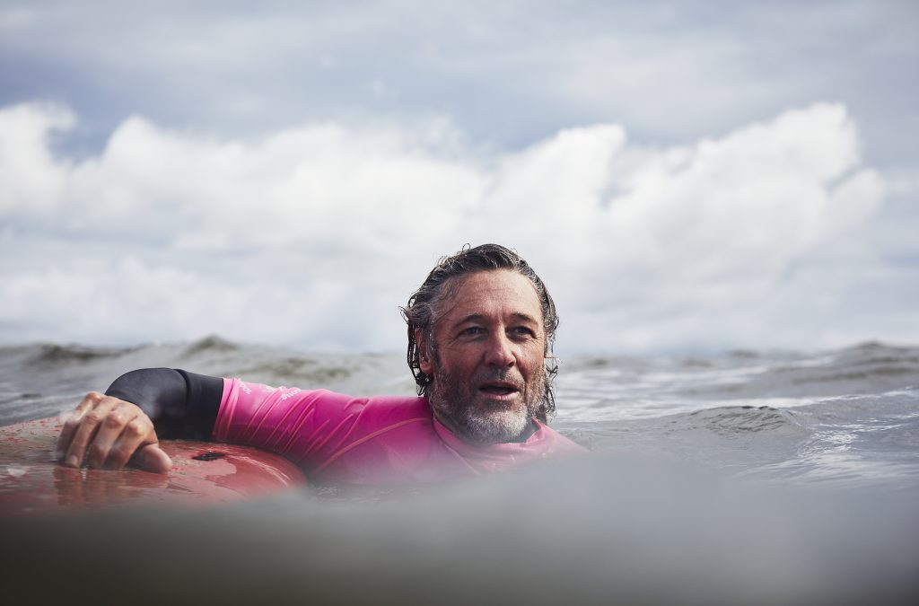 Adult male surfer with salt and pepper hair rests on surfboard in ocean water under cloudy sky, wearing pink rash guard.