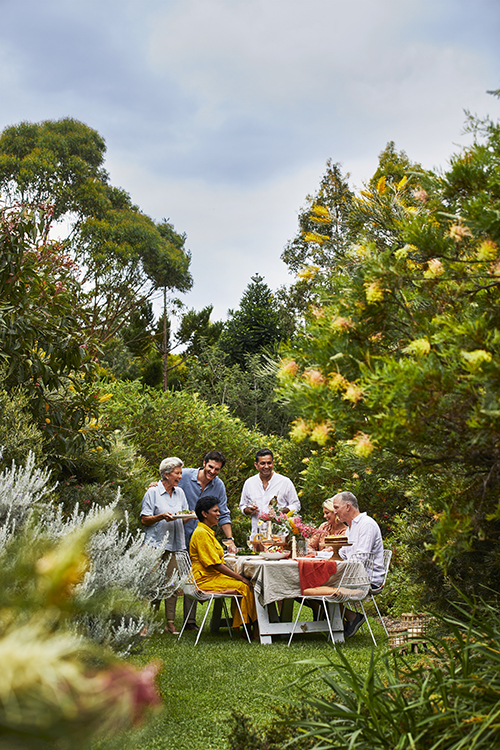A group of six people are enjoying an outdoor garden party, gathered around a table laden with food on a sunny day.