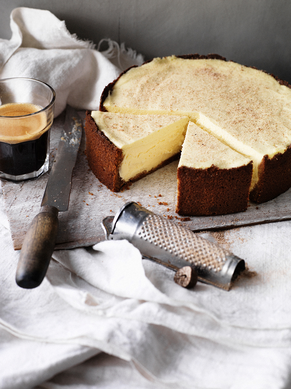Overhead shot of a cheesecake with a slice cut out, accompanied by a cup of coffee, a vintage grater, and a knife on a wooden surface.