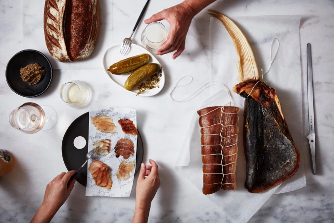 Overhead shot of food presentation with various dishes, including cured fish, pickles, bread, and beverages, on a white marble surface.