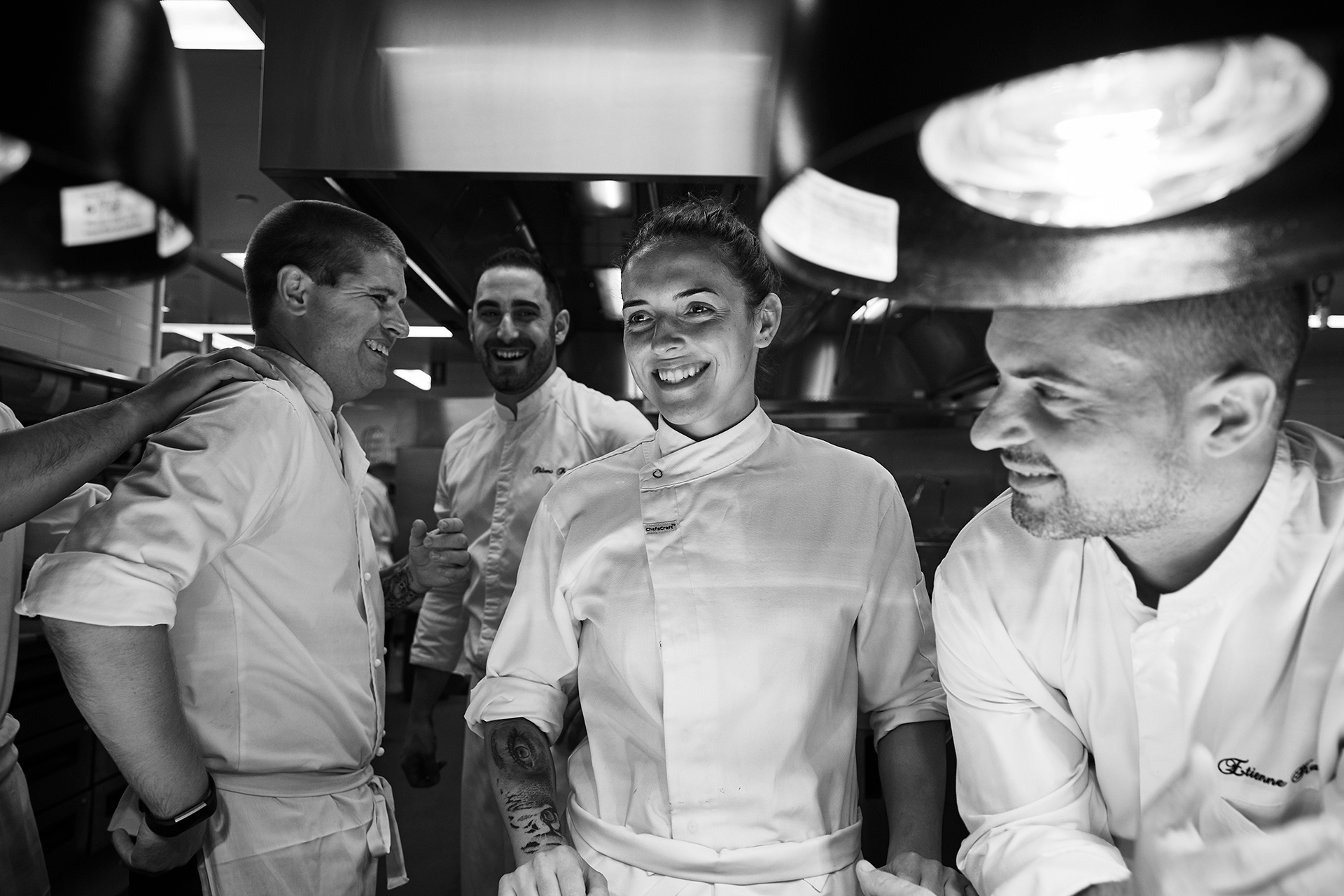Black and white shot of a group of chefs smiling in a commercial kitchen with metal fixtures and bright overhead lighting.