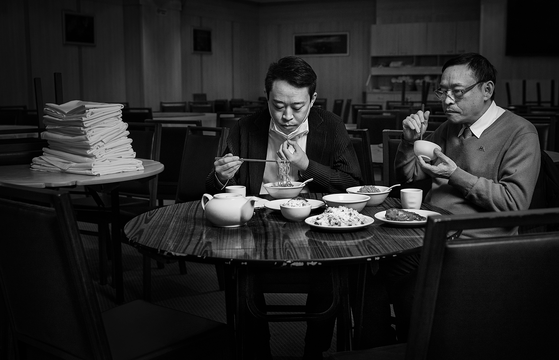 Black and white shot of two Asian men eating at a restaurant table laden with food, one wearing a mask under his chin.