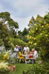 A group of six adults gathered around a dining table outdoors in a lush green garden setting, photographed by Rob Palmer.