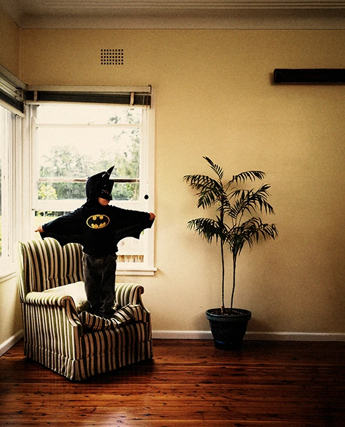 Child dressed in a Batman costume standing on an armchair inside a brightly lit room with a potted palm plant.