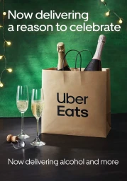 Brown paper delivery bag with Uber Eats logo containing two champagne bottles, flanked by two filled champagne glasses and a cork on a dark surface against a green backdrop with fairy lights