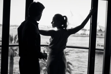 Silhouette of a man and woman holding wine glasses by a large window overlooking Sydney Harbour, photographed by Rob Palmer.