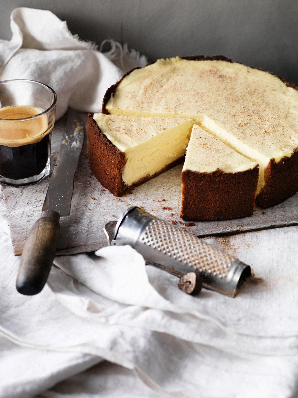 Whole baked cheesecake with a sliced wedge removed, set on a rustic surface with a glass of coffee and a small grater.