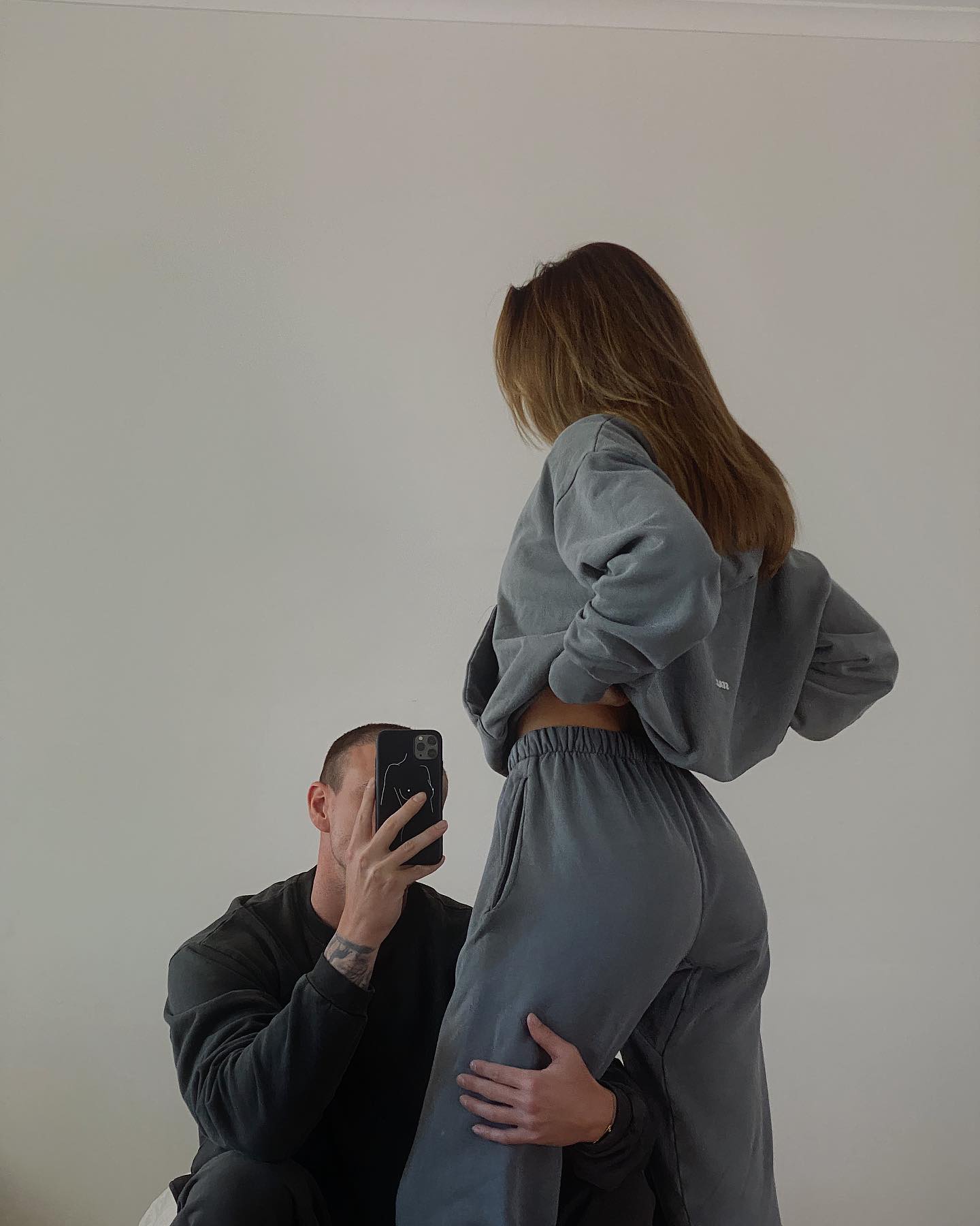 A man takes a photo of a woman wearing a grey sweatsuit in an indoor setting, capturing a casual and intimate moment with soft lighting.