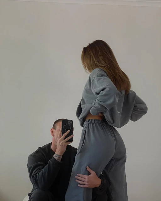 A man takes a photo of a woman wearing a grey sweatsuit in an indoor setting, capturing a casual and intimate moment with soft lighting.