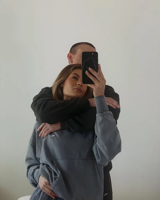 Woman in sweatshirt takes mirror selfie while embraced by man in black; casual fashion and indoor setting with soft natural light.