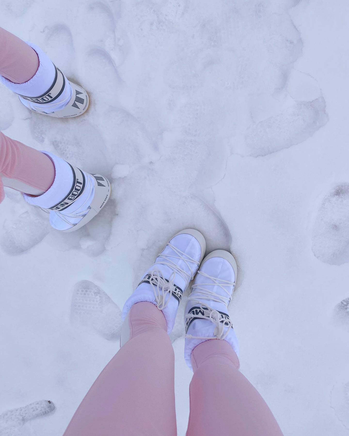 Two women stand in the snow wearing matching Moon Boot brand white winter boots and pink leggings, creating a bright, clean winter fashion statement.