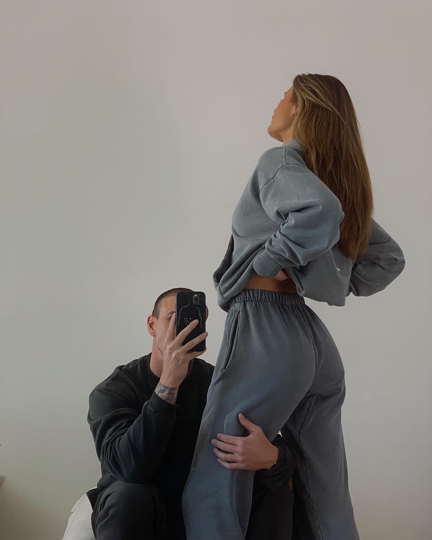 A man takes a picture of an adult female wearing a matching blue-grey sweatshirt and sweatpants in an indoor setting against a white wall.