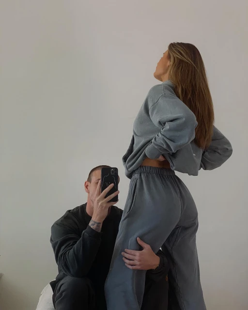 A man takes a picture of an adult female wearing a matching blue-grey sweatshirt and sweatpants in an indoor setting against a white wall.
