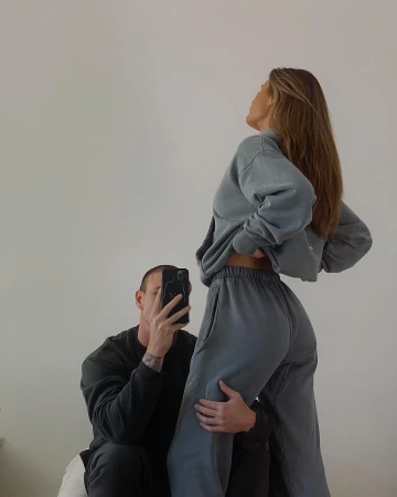 A man takes a picture of an adult female wearing a matching blue-grey sweatshirt and sweatpants in an indoor setting against a white wall.