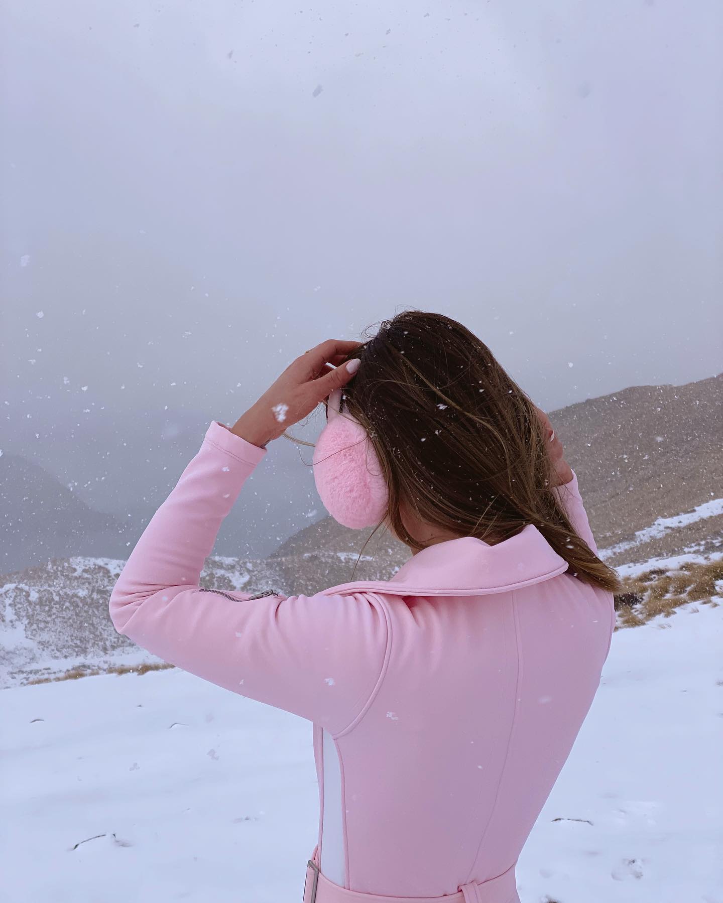 A young woman in pink ski suit and pink earmuffs stands in the snowy mountains on a cloudy day, looking out at the overcast landscape.