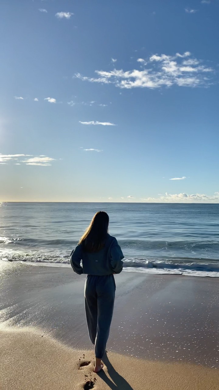 A woman walking on a sandy beach towards the ocean under a blue sky with scattered clouds during a sunny day with footprints visible.