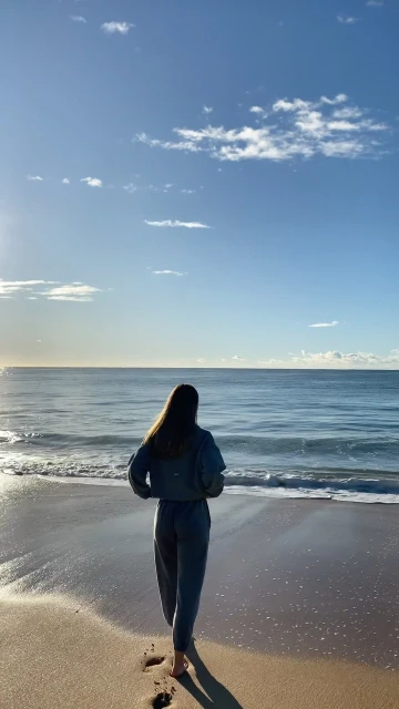 A woman walking on a sandy beach towards the ocean under a blue sky with scattered clouds during a sunny day with footprints visible.