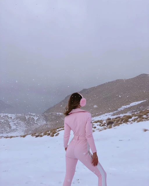 Woman in pink jumpsuit and earmuffs standing in snowy mountain landscape with light snowfall and overcast skies, viewed from behind.