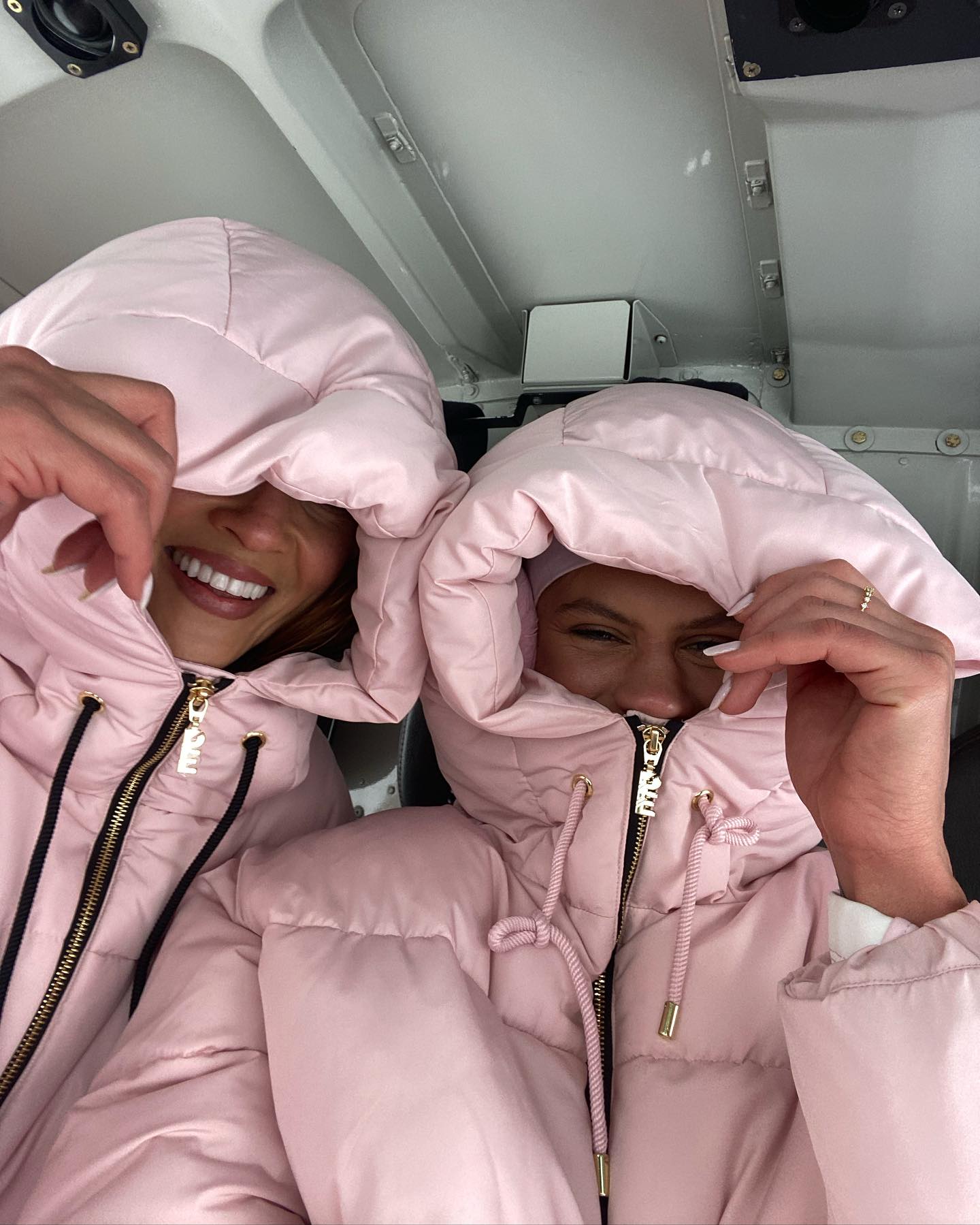 Two smiling women pose indoors, wearing matching pink puffer jackets with hoods up, partially obscuring their faces in a playful way.