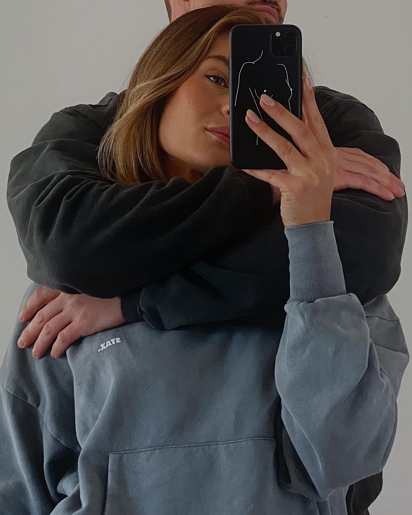 Woman takes selfie with phone case featuring line art of a female figure while a man embraces her from behind.