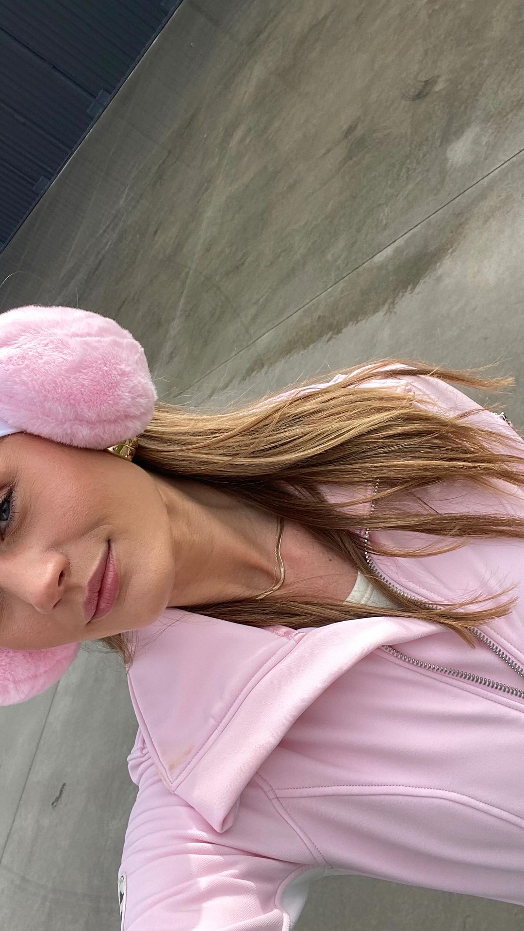 Portrait of a young woman wearing earmuffs and a pink jacket, posing indoors against a gray wall, capturing a modern and fashionable look.